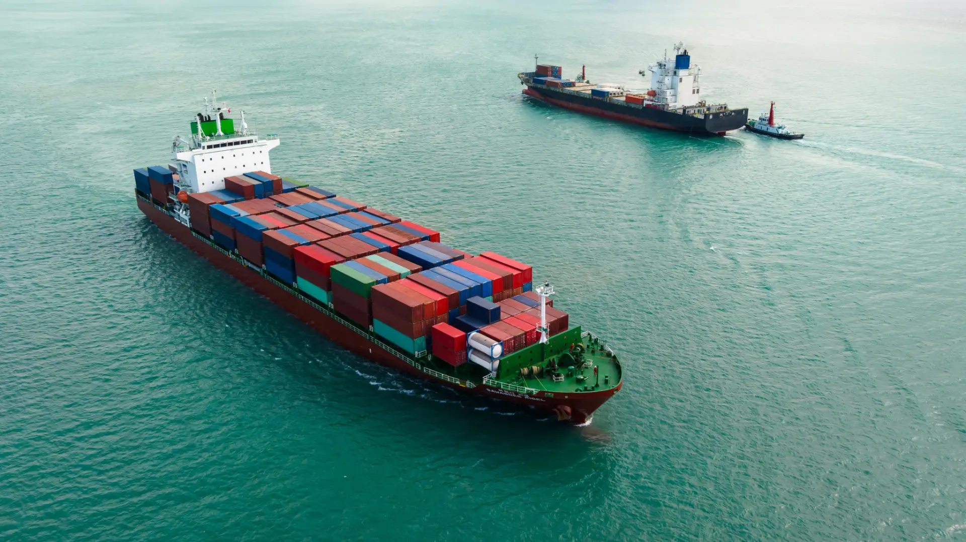 Aerial view of two container vessels and a tug manoeuvring in green coastal water.