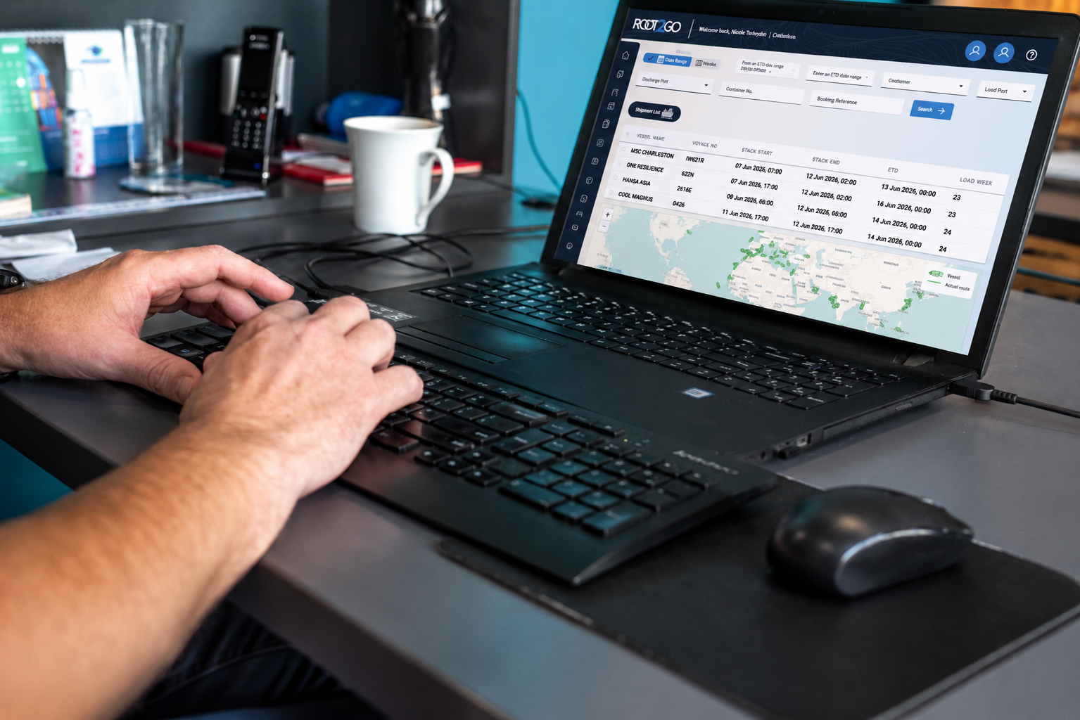 A laptop showing the Root2Go vessel schedule list and live shipment map on an office desk.